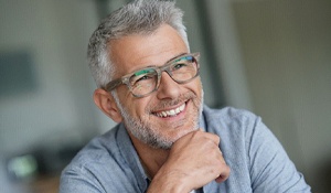 An older gentleman wearing glasses props his chin with his hand and smiles wide